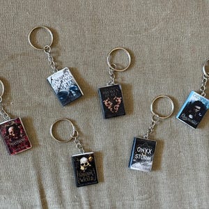 May include: Miniature book keychains with silver key rings. The keychains feature book covers with titles like "Serpent & Night", "Broken Heart", and "Onyx Storm". The keychains are on a tan fabric surface.