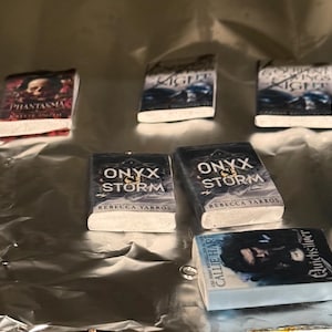 May include: Miniature book charms with various titles, including "Phantasma," "Onyx Storm," and "Kingdom of the Wicked." The book covers feature illustrations and text, with white pages and a silver background.