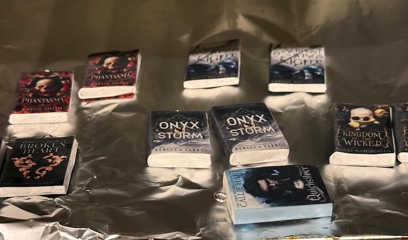May include: Miniature book charms with various titles, including "Phantasma," "Onyx Storm," and "Kingdom of the Wicked." The book covers feature illustrations and text, with white pages and a silver background.