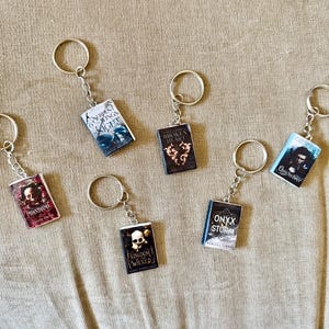 May include: A collection of miniature book keychains with silver key rings. The book covers feature various titles with colorful designs and text. The keychains are arranged on a tan surface.