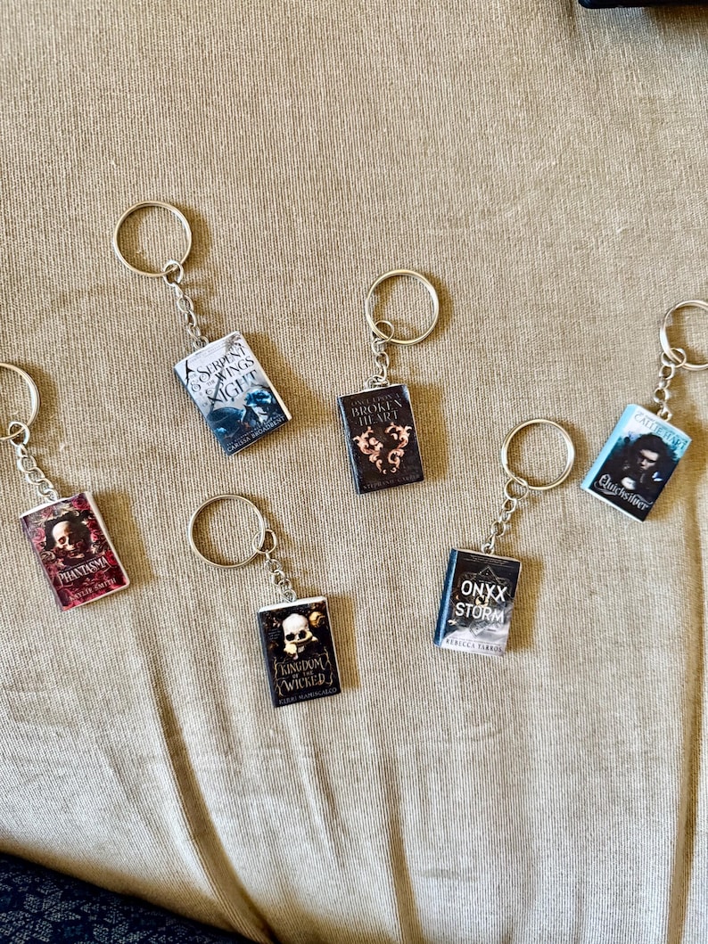 May include: A collection of miniature book keychains with silver key rings. The book covers feature various titles with colorful designs and text. The keychains are arranged on a tan surface.
