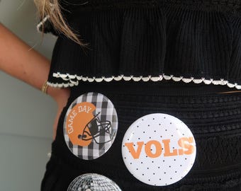 Könnte beinhalten: Schwarzes Strickoberteil und -rock mit weißem, gewelltem Besatz. Drei runde Knöpfe sind am Rock befestigt. Ein Knopf trägt die Aufschrift "GAME DAY" mit einer Footballhelm-Grafik. Ein anderer trägt die Aufschrift "VOLS" mit orangefarbenen Punkten. Der dritte Knopf trägt die Aufschrift "90 VOLS".