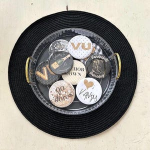 May include: A clear tray with gold handles holds several round buttons with various designs and text, including "VU", "Vandy", and "Go Dores". The tray sits on a black woven placemat.