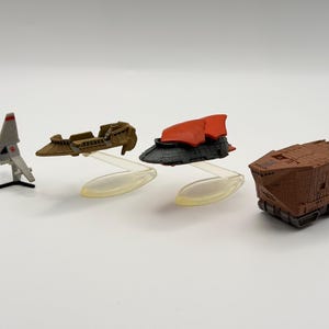 May include: A collection of miniature toy vehicles, including a silver and red rocket, a brown speeder, a black and red spaceship, and a brown cargo hauler. The toys are displayed on a white surface.