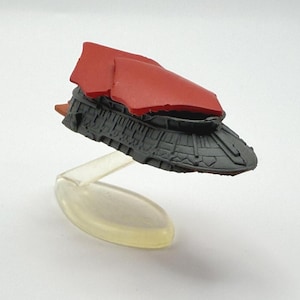 May include: A miniature spaceship model with a red top and a dark gray body. The model is suspended on a clear plastic stand, giving the impression of flight. The design features detailed textures and shapes, suggesting a futuristic aesthetic.