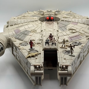 May include: A detailed model of the Millennium Falcon starship, featuring a light gray and beige color scheme with intricate paneling. Small figurines are positioned on the ship's surface, and the cockpit is visible. The model has a rectangular shape with a circular central section.