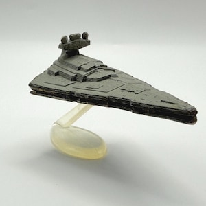 May include: A gray, miniature Star Destroyer model, a triangular spaceship with detailed surface features. The model is mounted on a clear, curved stand, and is set against a white background. The spaceship is approximately 6 inches long.