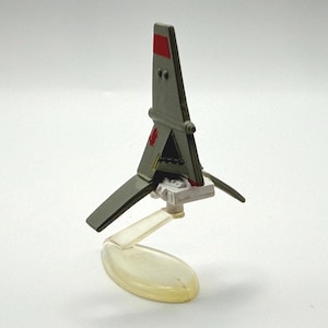 May include: A miniature gray and red spaceship model with angled wings and a clear plastic stand. The model has a detailed design with visible cockpit elements and is set against a plain white background.