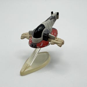 May include: A miniature toy spaceship, featuring a red, gray, and black color scheme. The model has wings and is mounted on a clear stand, suggesting a flying pose. The design is reminiscent of a science fiction vehicle.