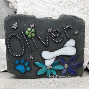 May include: A dark gray slate pet memorial stone with the name "Oliver" in silver-studded letters. Features a white bone, blue paw print, and colorful flowers. Includes the website "ChrisEmmert.com" and butterfly accents.