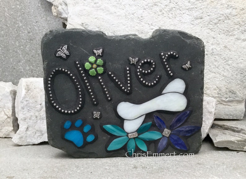 May include: A dark gray slate pet memorial stone with the name "Oliver" in silver-studded letters. Features a white bone, blue paw print, and colorful flowers. Includes the website "ChrisEmmert.com" and butterfly accents.
