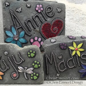 May include: Three personalized stone garden markers with names, flowers, paw prints, and butterfly accents. The names "Annie", "Juju", and "Madison" are displayed in a decorative font. The stones are gray and have a textured surface.