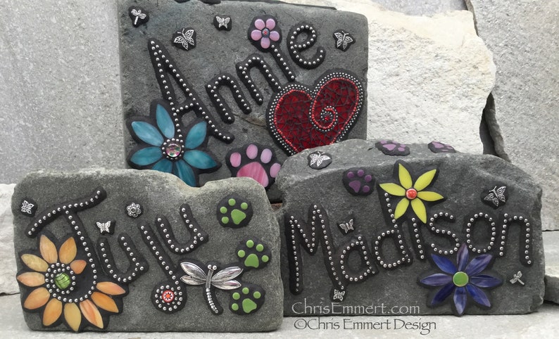 May include: Three personalized stone garden markers with names, flowers, paw prints, and butterfly accents. The names "Annie", "Juju", and "Madison" are displayed in a decorative font. The stones are gray and have a textured surface.