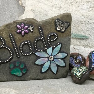 May include: A personalized stone garden marker with the name "Blade" in silver-dotted letters, adorned with a flower, paw print, and butterfly accents. Includes additional decorative heart-shaped stones.