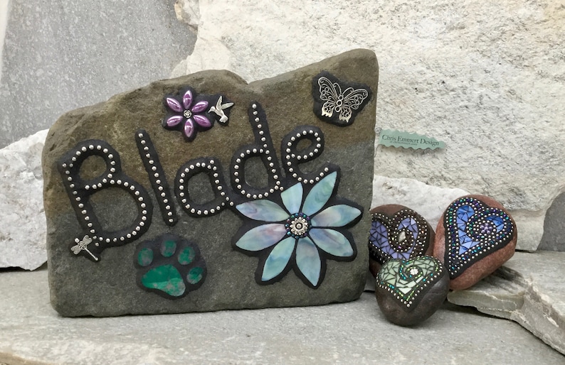 May include: A personalized stone garden marker with the name "Blade" in silver-dotted letters, adorned with a flower, paw print, and butterfly accents. Includes additional decorative heart-shaped stones.