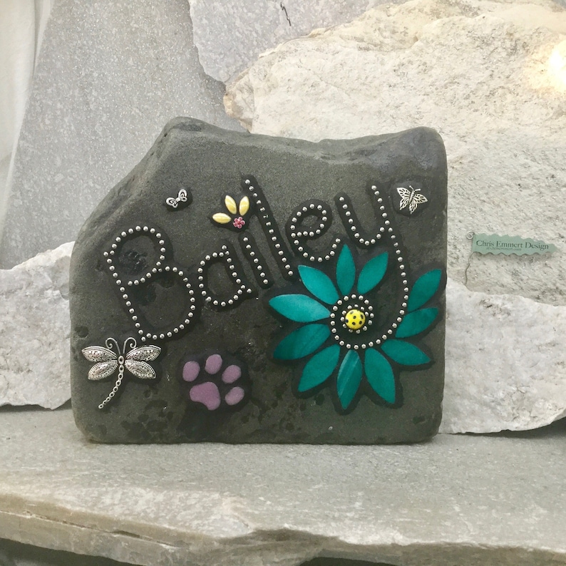 May include: A personalized stone garden marker with the name "Bailey" in silver-colored beaded letters. The dark gray stone features a teal flower, a purple paw print, and dragonfly and butterfly embellishments.