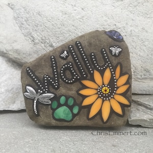 May include: A decorative stone with the name "Wally" spelled out in silver studs. The stone features a sunflower, a green paw print, and dragonfly and butterfly embellishments. The website address "ChrisEmmert.com" is visible.