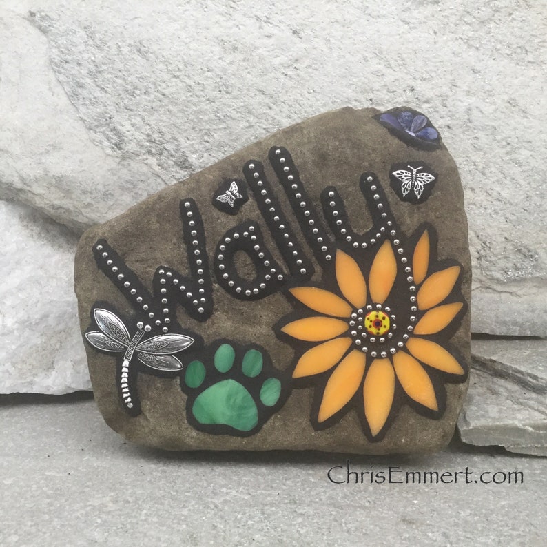 May include: A decorative stone with the name "Wally" spelled out in silver studs. The stone features a sunflower, a green paw print, and dragonfly and butterfly embellishments. The website address "ChrisEmmert.com" is visible.
