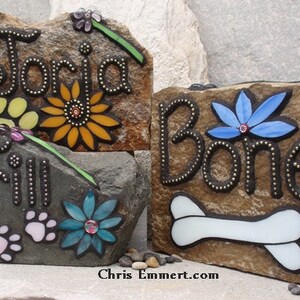 May include: Three decorative stone plaques with names and pet-themed designs. One reads "Toria" with a sunflower, another "Trill" with paw prints, and the last "Bone" with a bone and blue flower. The plaques are brown and gray.