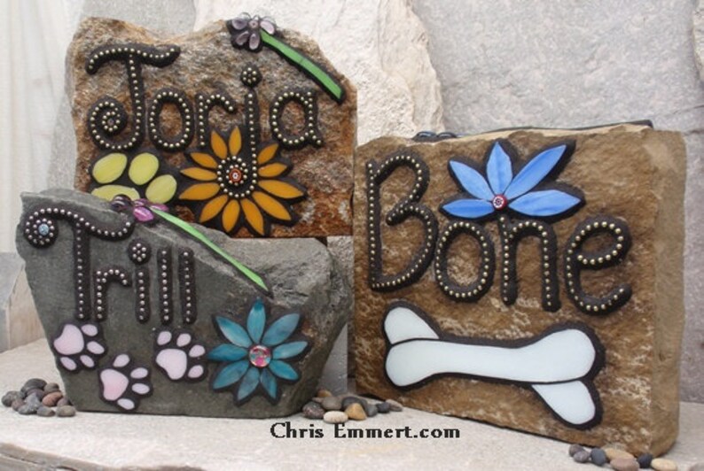 May include: Three decorative stone plaques with names and pet-themed designs. One reads "Toria" with a sunflower, another "Trill" with paw prints, and the last "Bone" with a bone and blue flower. The plaques are brown and gray.