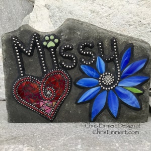 May include: A gray stone garden marker with the name "Missy" in black lettering. The design includes a red mosaic heart, a blue mosaic flower, and a paw print. The text "Chris Emmert Design at ChrisEmmert.com" is at the bottom.