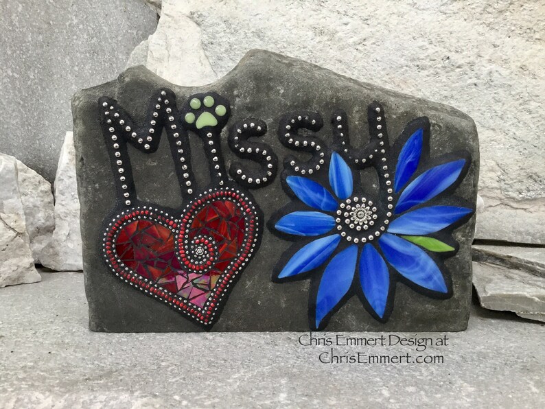May include: A gray stone garden marker with the name "Missy" in black lettering. The design includes a red mosaic heart, a blue mosaic flower, and a paw print. The text "Chris Emmert Design at ChrisEmmert.com" is at the bottom.