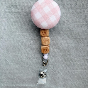 May include: A pink and white gingham badge reel with wooden letter beads spelling "SLP". The retractable reel has a clear plastic clip and a metal clasp. The badge reel is designed for professional use.