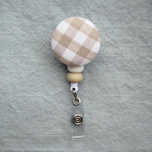 May include: A round badge reel with a tan and white gingham fabric-covered button. The reel has a clear plastic strap with a snap closure, and a series of neutral-colored beads. The background is a light blue-green fabric.
