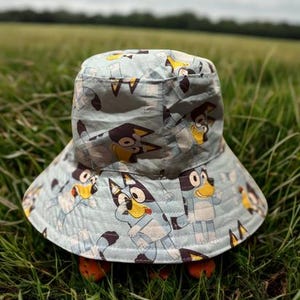 May include: A light blue bucket hat with a repeating cartoon dog design. The hat has a wide brim and is set in a field of green grass under a cloudy sky. The hat is designed for children.