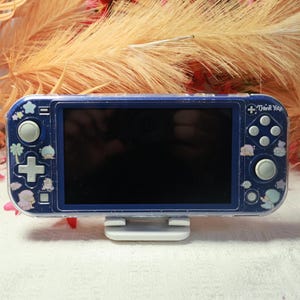 Peut inclure: Une console de jeu portable bleu foncé avec une coque de protection transparente à paillettes. L'appareil est orné de petites illustrations de personnages aux tons pastel, d'étoiles et de coquillages. Le texte "Thank You!" est inscrit en haut.