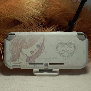 May include: A white handheld gaming console cover featuring a line art illustration of a person with long hair on the left side and a heart-shaped frame containing two rabbits on the right. The text "angelic doll" is printed in the centre.