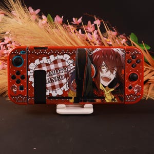 Puede incluir: Una funda roja para consola de juegos con un personaje de anime de cabello castaño y auriculares, un corazón blanco con patrón de cuadros rojos y el texto "ENOMOTO MINEO", "COLLAR" y "MALICE" impreso en la superficie del dispositivo.