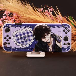 Puede incluir: Una funda para consola de juegos de color morado con un personaje de anime de cabello negro y auriculares. El diseño incluye un corazón a cuadros azul y blanco con el texto YANAGI AIJI, junto a iconos de personajes marcados COLLAR y MALICE.