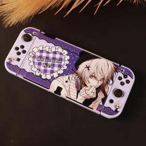 Puede incluir: Una consola de juegos portátil con una funda morada y blanca que presenta un patrón de corazón con volantes y el texto Okazaki Kei. El diseño incluye un personaje de estilo anime con cabello claro, auriculares y un gorro negro.