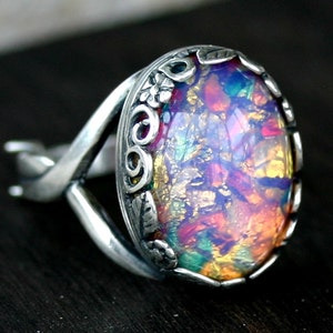 May include: A silver ring featuring an oval iridescent opal gemstone set in a detailed silver bezel. The bezel is decorated with intricate floral designs and swirls.
