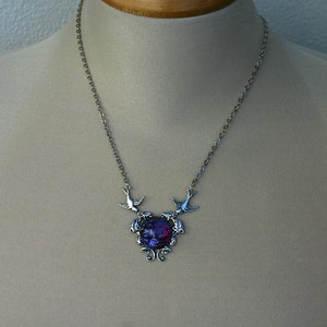 May include: Silver-toned necklace featuring a pendant with a vibrant, multi-colored center. Two bird-shaped charms flank the pendant, which is suspended from a delicate chain. The pendant has a swirling, abstract design.