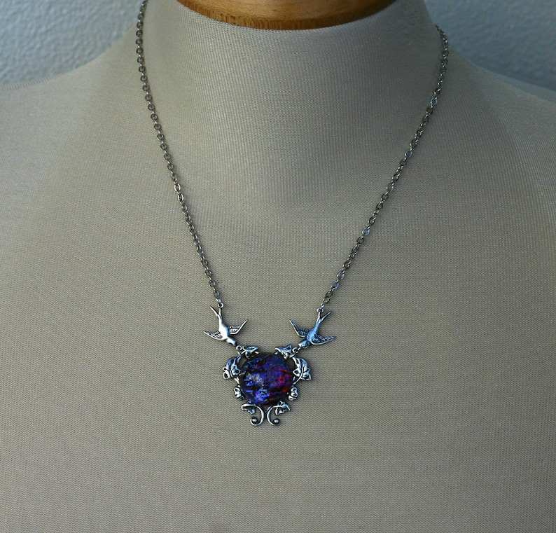 May include: Silver-toned necklace featuring a pendant with a vibrant, multi-colored center. Two bird-shaped charms flank the pendant, which is suspended from a delicate chain. The pendant has a swirling, abstract design.