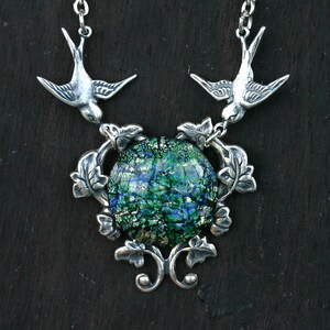 May include: Silver-toned necklace featuring two bird pendants flanking a central, round, dichroic glass pendant. The glass has a green and blue color scheme. The necklace has a detailed, nature-inspired design with leaf accents.