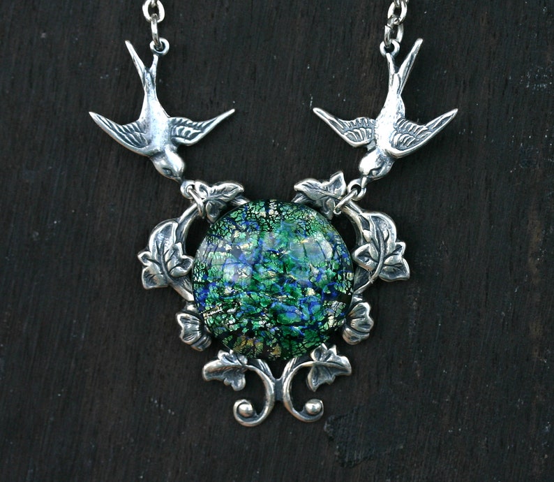 May include: Silver-toned necklace featuring two bird pendants flanking a central, round, dichroic glass pendant. The glass has a green and blue color scheme. The necklace has a detailed, nature-inspired design with leaf accents.