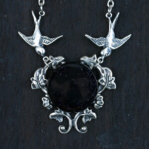 May include: A silver-toned necklace featuring a large, round, dark blue stone with sparkling inclusions. Two detailed bird pendants flank the stone, connected by ornate leaf and vine designs. The necklace has a delicate chain.