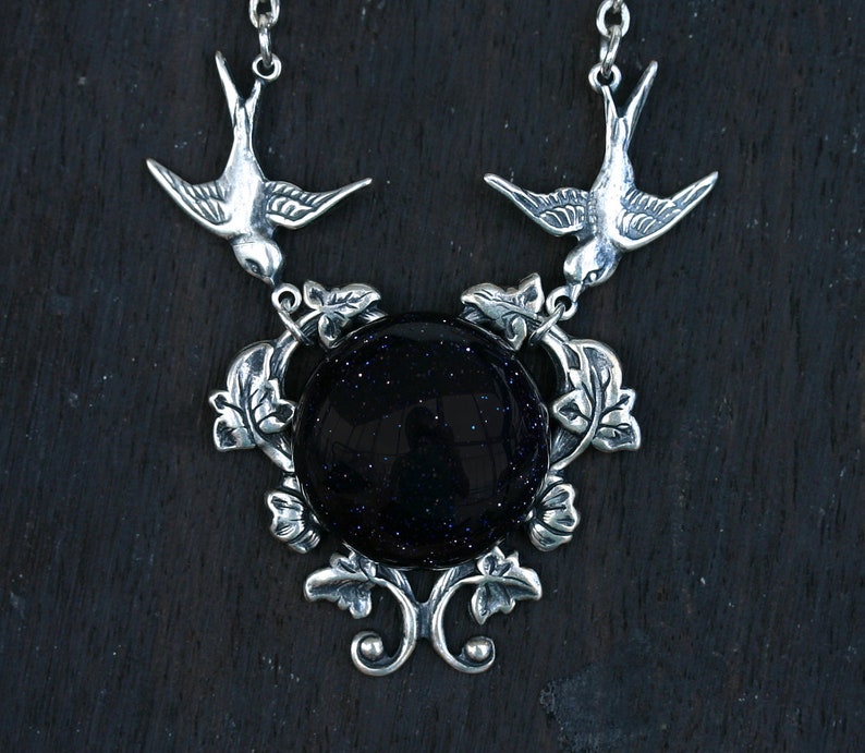 May include: A silver-toned necklace featuring a large, round, dark blue stone with sparkling inclusions. Two detailed bird pendants flank the stone, connected by ornate leaf and vine designs. The necklace has a delicate chain.