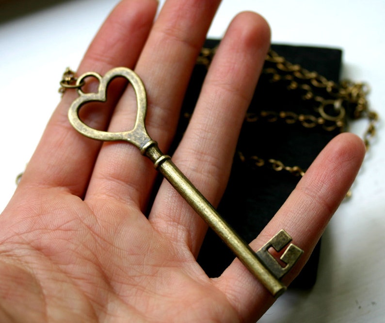 K&ouml;nnte beinhalten: Eine Halskette mit einem Anh&auml;nger in Form eines Schl&uuml;ssels aus Messing im Vintage-Stil. Der Schl&uuml;ssel hat eine herzf&ouml;rmige Spitze und einen rechteckigen Bart. Die Kette ist messingfarben. Ein romantisches Accessoire.