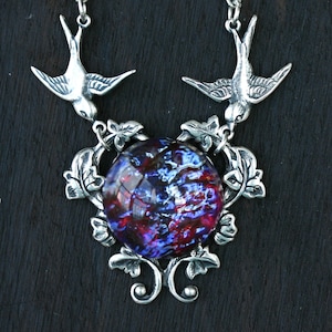 May include: Silver-toned pendant necklace featuring two birds flanking a large, round, dichroic glass stone. The stone displays a mix of blue, red, and purple hues. The pendant is adorned with leaf and vine details, creating an elegant, nature-inspired design.