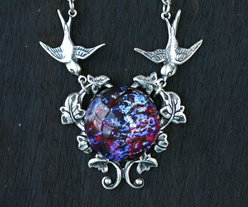 May include: Silver-toned pendant necklace featuring two birds flanking a large, round, dichroic glass stone. The stone displays a mix of blue, red, and purple hues. The pendant is adorned with leaf and vine details, creating an elegant, nature-inspired design.