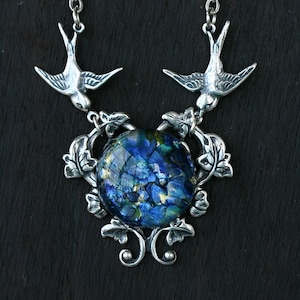 May include: Silver-toned necklace featuring two bird pendants flanking a large, round, dichroic glass pendant. The glass has a blue and gold floral pattern. The necklace has a decorative floral and leaf design around the glass pendant.