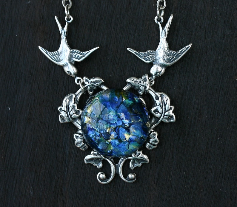 May include: Silver-toned necklace featuring two bird pendants flanking a large, round, dichroic glass pendant. The glass has a blue and gold floral pattern. The necklace has a decorative floral and leaf design around the glass pendant.