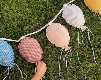 Peut inclure: Une guirlande décorative composée de cinq ballons en tissu rembourré aux motifs et couleurs variés, incluant des lignes ondulées bleues, un vichy rouge, un rose uni, des rayures blanches et grises, et un tissu jaune texturé sur un ruban blanc.