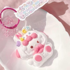 May include: A white, bunny-shaped keychain with pink accents, a pink bow, and a pink keychain ring. The keychain is on a white, leaf-shaped dish. A tag with the text "Pawkemons" is attached to the keychain.