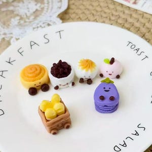 May include: A white plate with the words "BREAKFAST", "TOAST", "COFFEE", "SALAD", and "FRUIT" in black lettering. The plate holds six miniature food items, including a cinnamon roll, a bowl of dark berries, and a purple macaron.