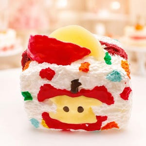 May include: A decorative, square-shaped cake with white frosting, topped with a red apple slice and a yellow fruit. The cake features a red apple design on the front, with colorful sprinkles. The overall theme is a whimsical dessert.